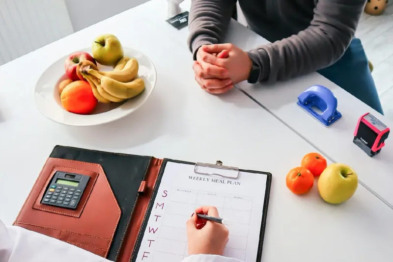 Two people at a table writing a weekly meal plan on a clipboard, beside a bowl of fruit